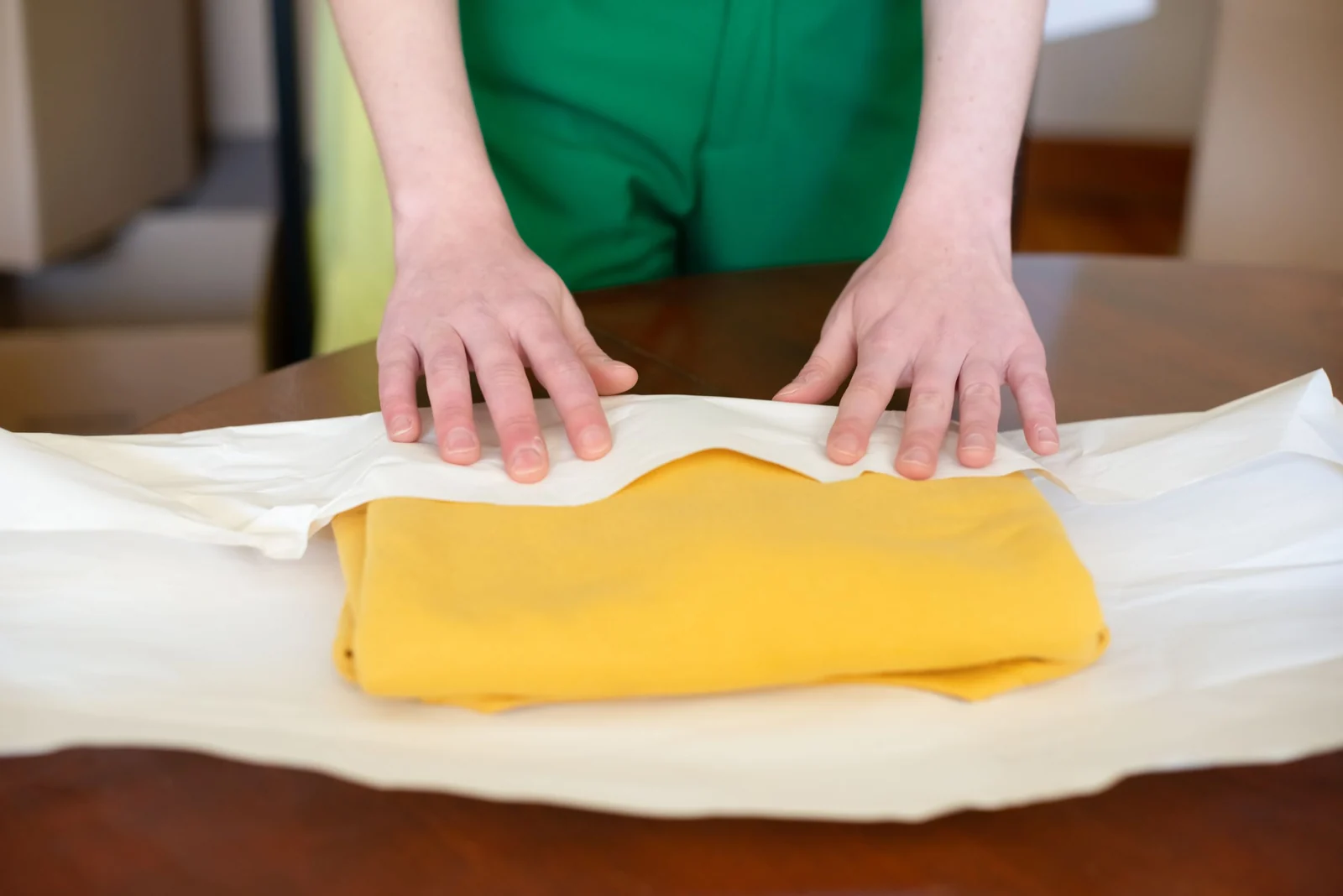 Hands wrapping a folded yellow garment in tissue paper.